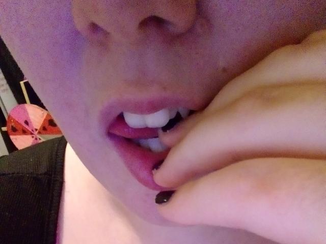 LoveClos Gesicht zeigt einen leicht rosafarbenen Teint, wobei ihre Zunge leicht herausragt und sie an der dunklen Nagel ihres Fingers knabbert. Ihre weißen Zähne sind sichtbar. LoveClos Hand liegt auf ihrem Kinn..