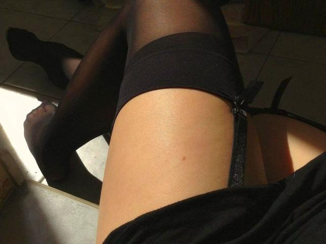 LadyMiller's thighs in black sheer stockings with garter straps, showing a sunlit, slightly shiny butt, her legs crossed, and the floor in the background.
