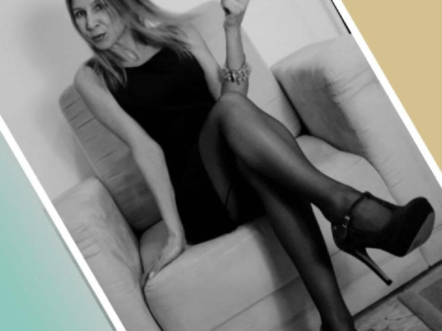 KatarinaDamo sitzt in einem Stuhl, trägt ein schwarzes Kleid, feine Strumpfhosen und High Heels, wobei sie ihre Beine zur Schau stellt und eine Zigarette hält. Ihre Brüste sind klein..