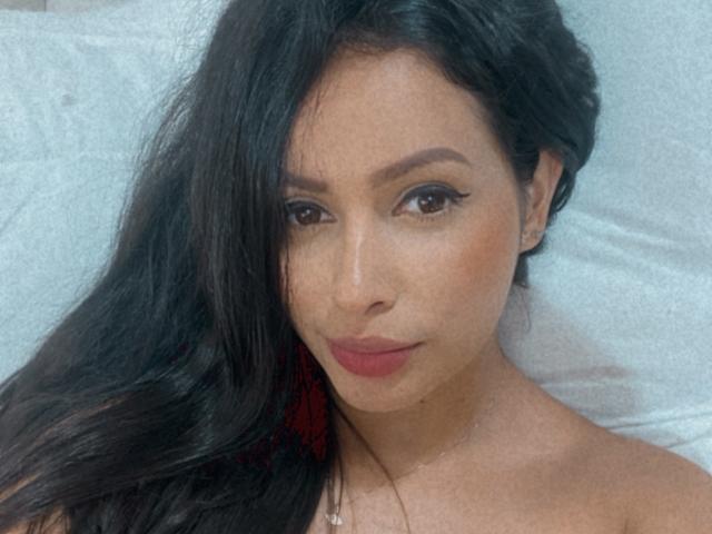 EmilyParkerr has long black hair, brown eyes, and red lipstick. She's looking at the camera with a slight smile, wearing a necklace, and has a light brown skin tone.