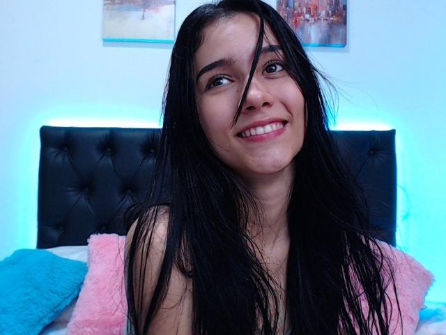 DeisyWest has long black hair, a cute smile, and is showing her pearly whites. She's topless with a cute pink and blue pillow behind her.
