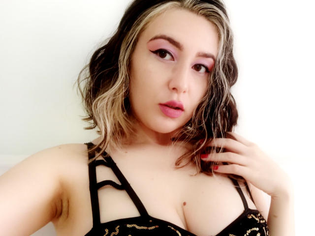 MabyLegrand with blonde and brown wavy hair, pink eyeshadow, and pink lipstick, wearing a black strappy top, showing her tits, with a hand near her face.