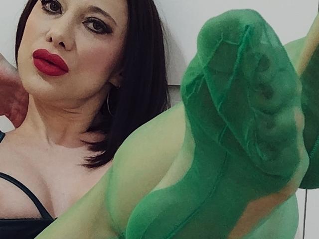WandaDominatrix avec du rouge à lèvres, un soutien-gorge noir et des bas verts transparents, montrant son pied, la plante visible, dans un plan légèrement incliné. Ses seins sont petits, son visage est sérieux..