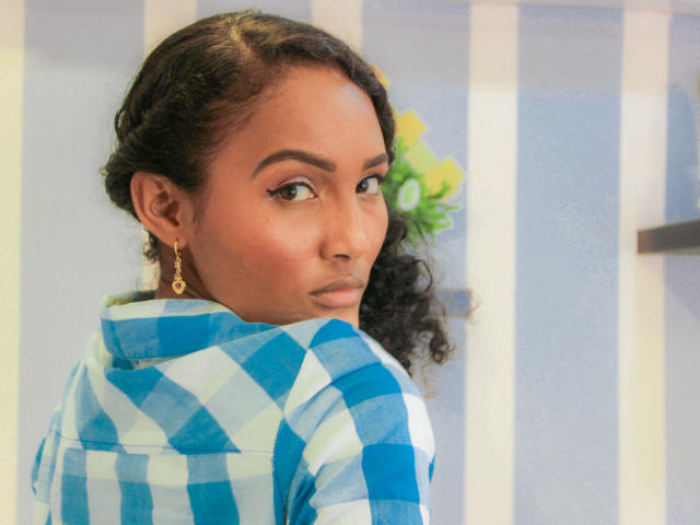 Megan Gil, avec ses cheveux noirs bouclés, portant une chemise à carreaux bleu et blanc, des boucles d'oreilles dorées et un maquillage discret, regardant par-dessus son épaule avec une expression sérieuse..