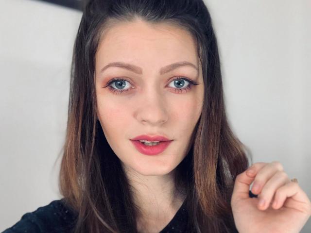 BlossomPussy hat blaue Augen, roten Lippenstift und braunes, welliges Haar, mit einem leichten Lächeln, und schaut direkt in die Kamera. Sie trägt ein schwarzes Oberteil und hält ihr Haar..