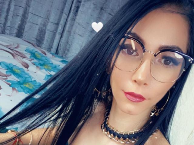 Crazysin has long black hair, wearing big glasses, gold earrings, and a black choker with gold details. She's got red lipstick and a white heart above her head.