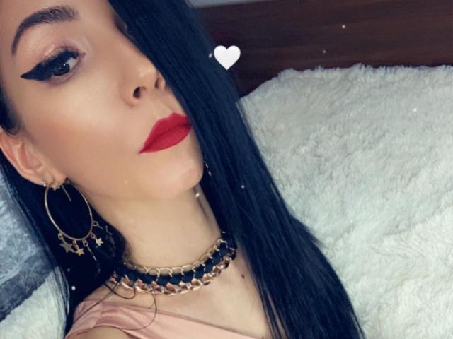 Een gekke vrouw met lang zwart haar, rode lippenstift, opvallende eyeliner, gouden oorringen, een sterrenketting, een roze top, liggend op een wit, pluizig oppervlak, met een klein hartje-emoji boven haar hoofd..