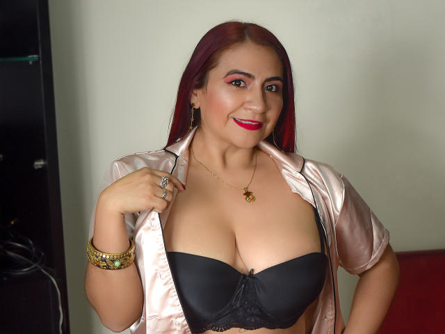 NahelaBigMelonsX with red hair, smiling, wearing a shiny pink satin shirt, black bra, showing big tits, red lipstick, gold bracelet, necklace, holding shirt open.