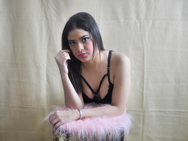 DulcesMiel with long dark hair, pink lipstick, black strappy lingerie, leaning on a pink furry stool, looking at camera with right hand touching chin, beige backdrop.