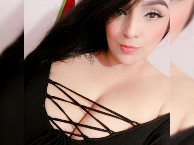 Briana69x con capelli neri, guance rosate e una maglietta nera con lacci che mette in mostra il suo seno abbondante. Tatuaggio sulla spalla sinistra. Sorriso dolce, occhi marroni, trucco leggero..