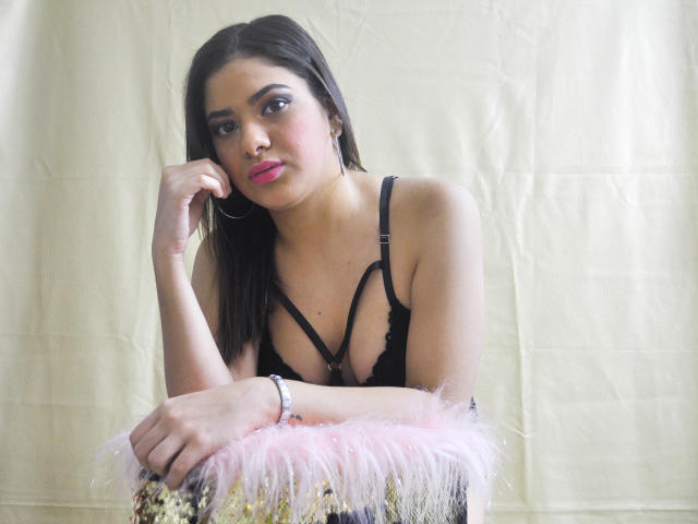 DulcesMiel with dark brown hair, pink lipstick, black lace bra, and pink furry stool, resting chin on hand, looking at camera, wearing silver bracelet.