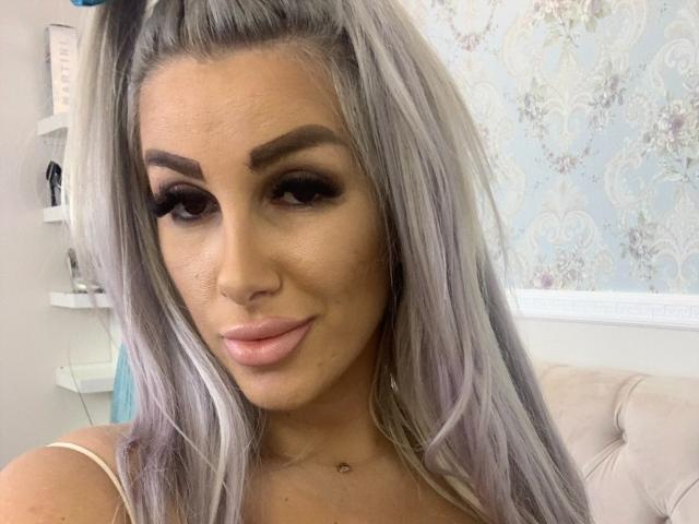 TanyaMonroe with long, silver-gray hair, dark eyebrows, and pink lips, wearing a white top, showing a cute, slightly smirking face. Her tits are small and she has a nose ring.