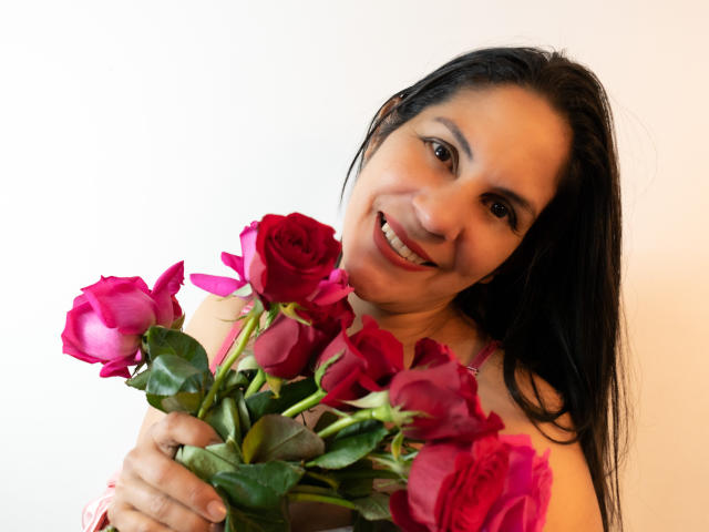 RoraimAnaggie with dark hair, smiling, holding a bouquet of red roses, wearing a pink top, looking at the camera, showing cute tits, and a cute ass.