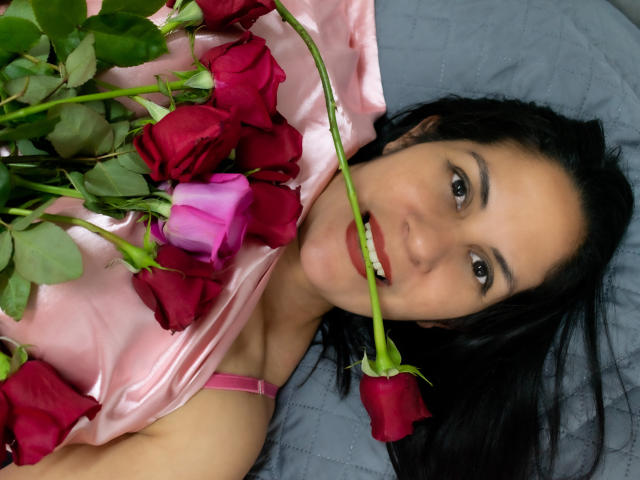 RoraimAnaggie, com cabelo escuro, vestido rosa, deitada em uma cama cinza, segurando rosas vermelhas e rosas, uma rosa na boca, olhando para a câmera com batom vermelho, olhos verdes..