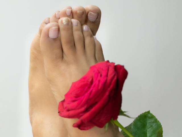 RoraimAnaggie's foot with pink nail polish, holding a red rose. Rose's green leaf visible, RoraimAnaggie's toes pointed upward. Simple, clean background.