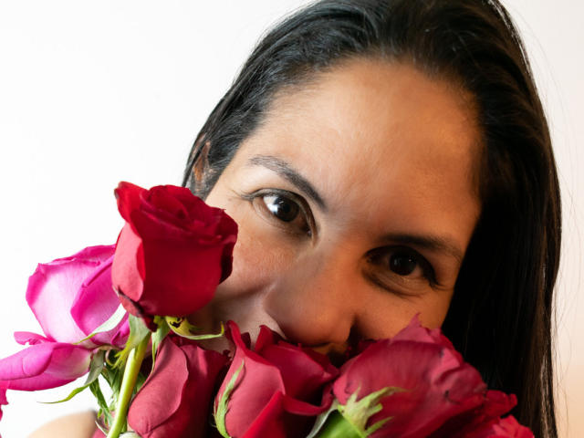 Das Gesicht von RoraimAnaggie ist teilweise von einem Strauß roter Rosen verdeckt, ihre dunklen Augen blicken mit einem leichten Lächeln nach oben, ihr schwarzes Haar fällt zur Seite..