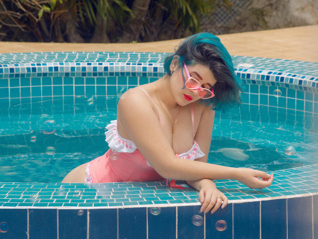 MaraMaison, con i capelli color acquamarina, indossa un costume intero rosa con volant e occhiali da sole rosa, appoggiata al bordo della piscina piastrellata di blu, con l'acqua fino al petto, guarda in basso, con rossetto rosa, sexy, di origine asiatica..