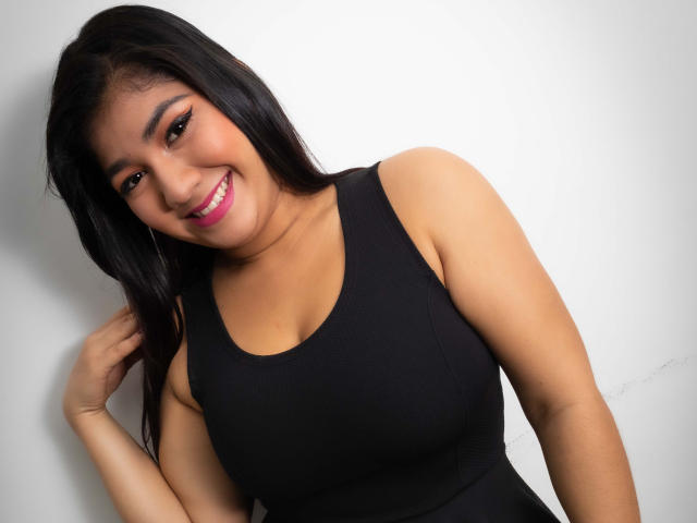 Kendall Owens, com longos cabelos pretos, sorrindo, batom rosa, top preto, mostrando os seios, em frente a uma parede branca, com a mão tocando o cabelo, parecendo linda e sexy..