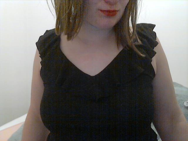 VraieFrancaise with shoulder-length brown hair, wearing a black ruffled sleeve top, showing her large tits, slightly visible cleavage, and red lipstick.