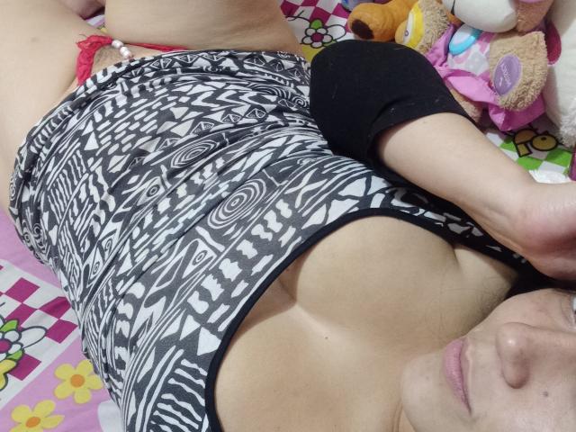 HotNastycamila lying on bed, wearing black and white patterned top, exposing small tits, with red lace panties, black sleeves, and stuffed toys around. Her face is partially visible.