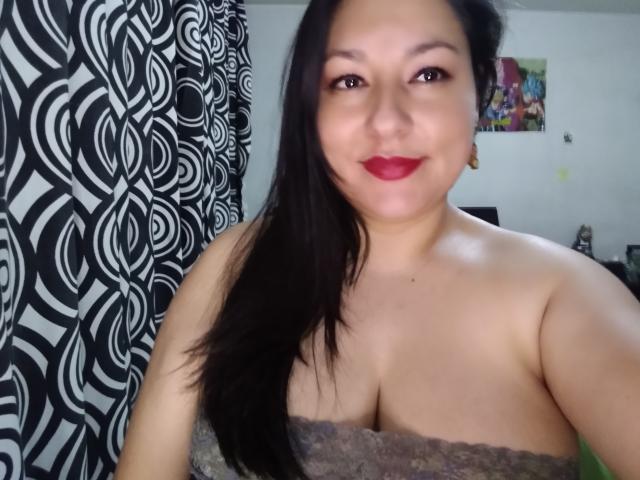 SakotoDika with black hair, red lipstick, and gray lace top showing her tits, smiling at the camera, in a room with a black-and-white patterned curtain.