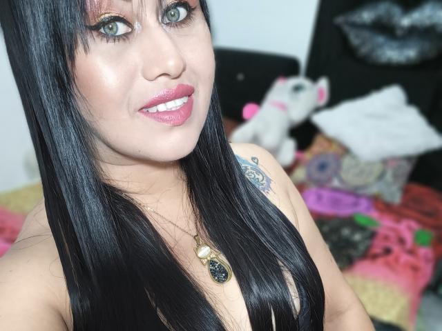 CathyCat hat schwarzes Haar, grüne Augen, pinken Lippenstift und ein Tattoo an ihrer rechten Schulter. Sie trägt eine Halskette mit einem schwarzen Anhänger, die ihre Brüste zeigt..