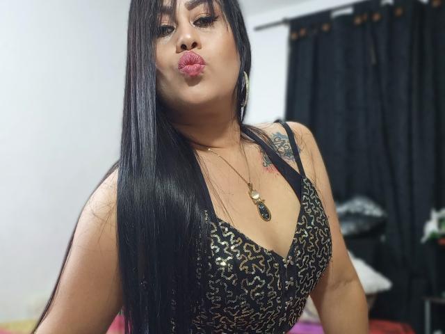 CathyCat, avec de longs cheveux noirs, des lèvres roses pincées, portant une robe à léopard, montrant ses seins, un tatouage sur l'épaule droite, un collier en or, des rideaux noirs en arrière-plan..