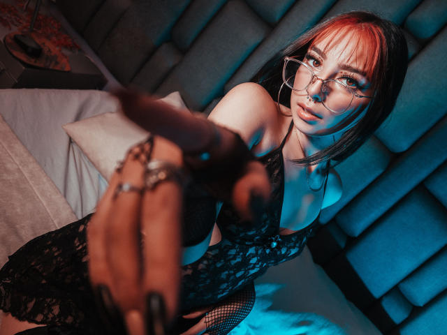 ConnieLuna, con capelli rossi e neri, occhiali e biancheria intima in pizzo, che indica in avanti, rivelando calze a rete nere e un anello sulla mano destra, in una camera da letto scarsamente illuminata..
