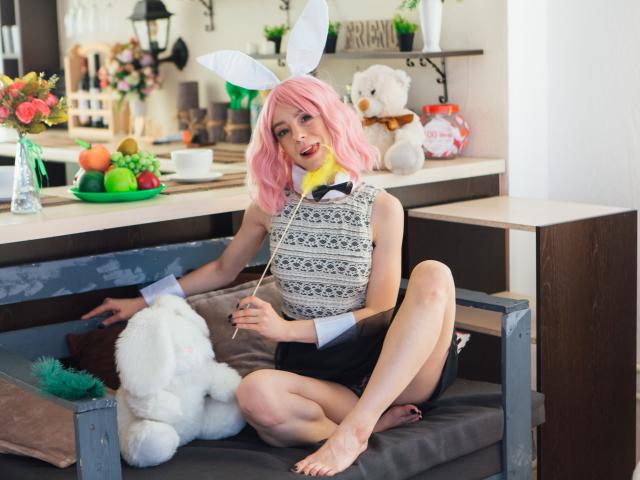 MissAstravert with pink hair, white bunny ears, and a grey dress sits on a bench, legs crossed, holding a white string, flirty smile, black skirt, stuffed bunny beside her.