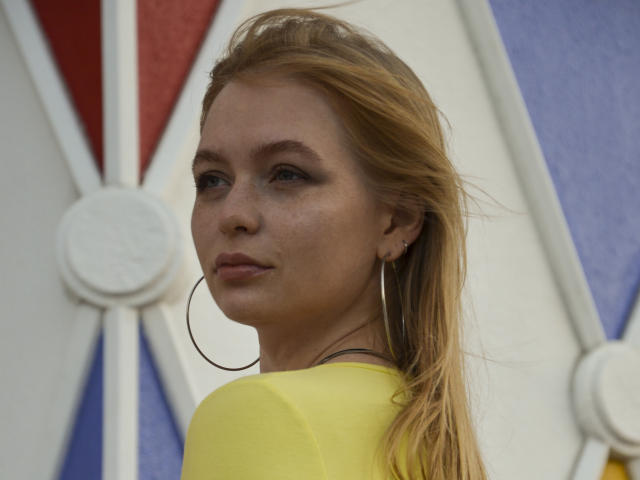 SweetAleksaX with blonde hair, hoop earrings, and freckles, wearing a yellow top, looking back with a slight smile, showing her ass in the photo.
