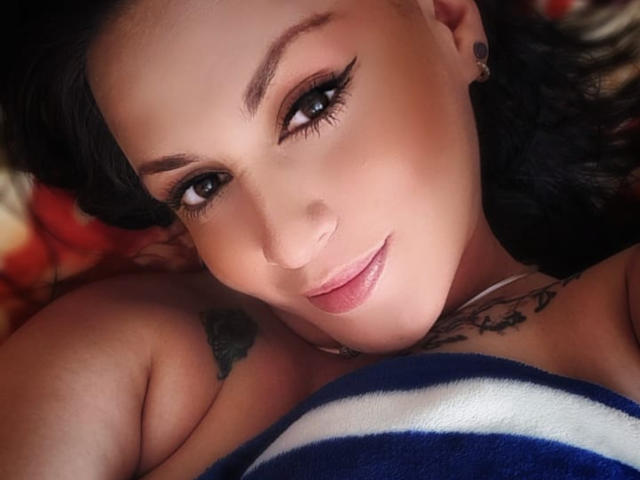 ElinaJhonson with dark hair, black eyeliner, and pink lipstick, lying down, smiling, showing tattoos on her shoulder and chest, with a blue and white striped blanket.