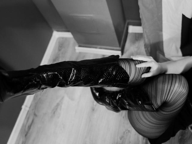 As brilhantes botas pretas de cano alto da Radiancee, com textura de padrão de pele de cobra, sendo delicadamente tocadas por sua mão, sentada em um chão de madeira, em uma foto em preto e branco..