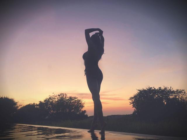 Silueta de CurlyAmeli con el cabello rizado, con los brazos levantados, de pie sobre una superficie húmeda, atardecer en el fondo, cabello largo, cuerpo curvilíneo, senos pequeños, delgada, sexy, agua reflectante..