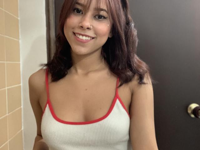 Annie Roberts, sorrindo com cabelos castanhos ondulados, veste uma regata branca texturizada com alças vermelhas, que revelam seios pequenos, e usa um piercing no nariz, estando em um banheiro..