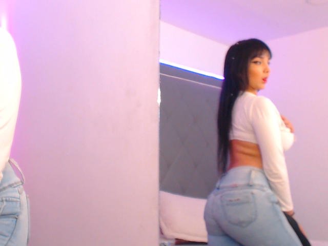TamaraOlsen with long black hair, white crop top, and blue jeans, showing her ass in mirror reflection, looking back with lips slightly parted.