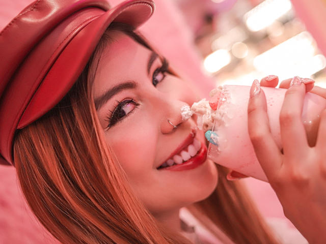 IsaTimeSweet with red hat, pink nails, nose ring, eating pink ice cream, smiling, showing teeth, pink lipstick, blonde hair, bokeh background, holding ice cream with white sprinkles.