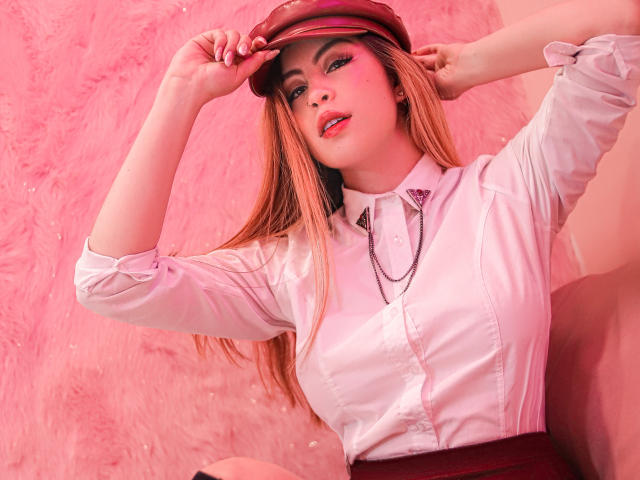IsaTimeSweet with pink lipstick, wearing a white shirt, red hat, and necklace, touching her hat, showing her tits, pink makeup, and long blonde hair, in a pink setting.
