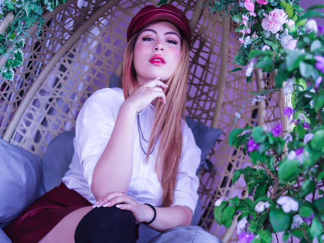 IsaTimeSweet, avec de longs cheveux blonds, un chapeau rouge, une chemise blanche, une jupe rouge, des bas noirs montants jusqu'à la cuisse, des lèvres pulpeuses, un doigt sur la joue, entourée de fleurs, assise dans un fauteuil en osier..