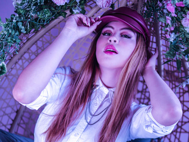 IsaTimeSweet with long brown hair, pink lipstick, wearing a maroon cap, white shirt, and necklace, adjusting her cap with both hands, looking seductive.