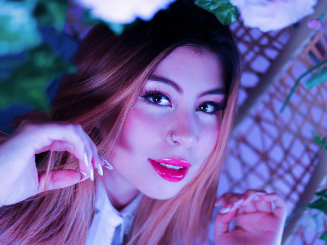 IsaTimeSweet with pink lipstick, blue eyes, and a nose ring, has long pinkish-brown hair, white nails, and a sexy look, holding hair near her face.