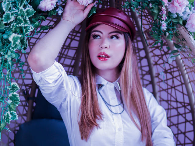 IsaTimeSweet, con cabello castaño largo, gorro rojo, camisa blanca y collar, mirando hacia arriba con una mano en las flores, tiene labios rosados brillantes y una cara linda, mostrando sus pechos..