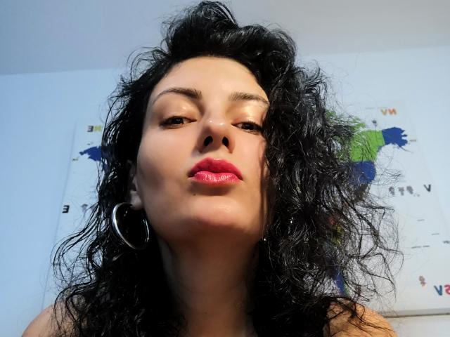 AddictedToUTS with curly black hair, shiny skin, red lipstick, and hoop earrings, looking up at the camera with a slightly pouty expression. Puffy nose, subtle makeup.