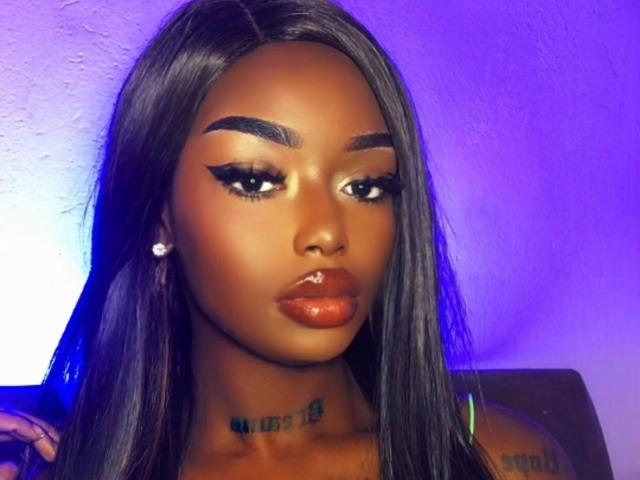 EbonyQueeny has dark brown skin, long straight black hair, thick eyebrows, winged eyeliner, shiny orange lipstick, and a small diamond earring. Her chest tattoo is visible.