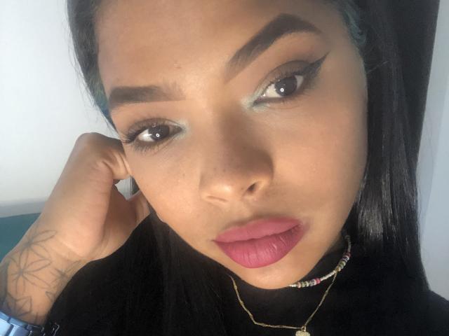 VickyGreys with dark hair, brown eyes, and pink lipstick, wearing bold eyeliner, touching her ear, black shirt, gold necklace, and tattoo on her wrist.