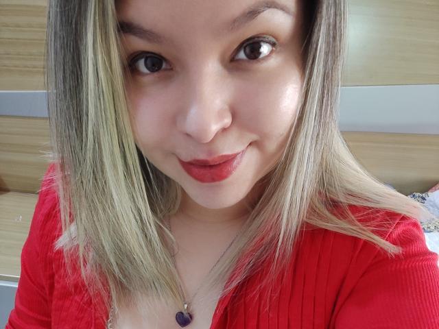 Myalewwis heeft een lichtbruine huid, blond steil haar en rode lippenstift, draagt een rode cardigan met een paarse ketting en glimlacht naar de camera..