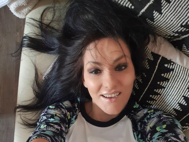 JoliDanielle's face, smiling with dark hair spread out, wearing a white and black floral raglan shirt, lying on a patterned pillow, looking up at the camera.