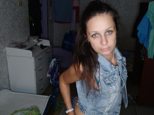 Emily Rubie, con lunghi capelli castani e occhi azzurri, indossa un gilet di jeans senza maniche, guarda la fotocamera, è in piedi in una stanza disordinata, mostrando la schiena nuda e un braccialetto bianco..