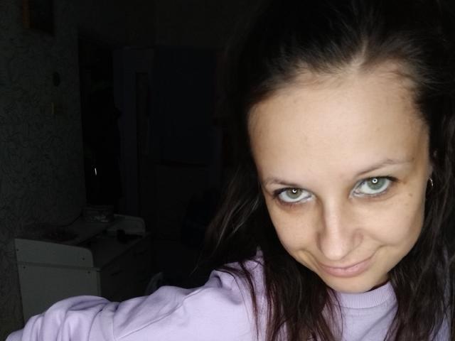 Emily Rubie, mit grünen Augen, die leicht lächelt, trägt einen violetten Pullover, hat dunkelbraunes Haar, helle Haut und ein dezentes Nasenpiercing. Ihre Brüste sind nicht sichtbar..