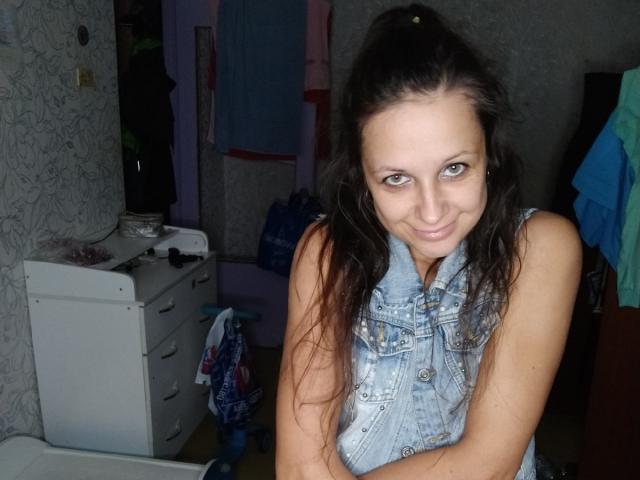 Emily Rubie, con lunghi capelli castani, occhi azzurri e un gilet di jeans senza maniche, sorride alla fotocamera, con le braccia incrociate, mostrando un viso carino e attraente con piccoli seni..