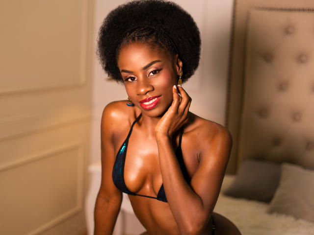 MiaHopson with dark skin, afro, red lipstick, wearing a black bikini, hand on cheek, smiling, small but perky tits, sexy, in a bedroom with a tufted headboard.